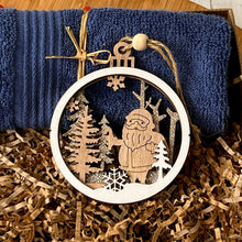 Load image into Gallery viewer, Close-up of a laser-cut wooden Christmas ornament featuring a Santa figure, snowflake, and glittered trees, resting on shredded paper with a navy washcloth in the background. Part of a festive menβs self care gift box.
