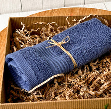Load image into Gallery viewer, Rolled navy blue cotton washcloth tied with natural raffia, nestled in kraft paper shred inside a gift box. A part of a winter self care package for men with a cozy and minimalist aesthetic.