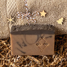 Load image into Gallery viewer, Handcrafted brown soap bar with a dark textured top and subtle black swirls, embossed with a geometric design, displayed on kraft paper filler in front of a folded beige towel decorated with small wooden holiday shapes.