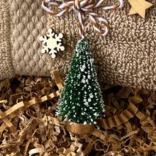 Load image into Gallery viewer, Miniature green Christmas tree ornament with a wooden base, dusted with artificial snow, displayed on kraft paper filler in front of a folded beige textured towel decorated with small wooden holiday shapes.