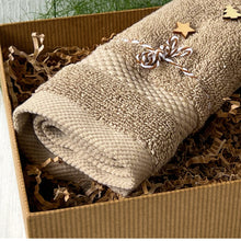 Load image into Gallery viewer, Rolled beige textured towel tied with a white and brown twine bow, decorated with small wooden star and tree shapes, placed on kraft paper filler inside a gift box.