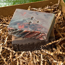 Load image into Gallery viewer, Two handcrafted soap bars stacked on kraft paper filler: the top bar features a marbled gray, red, and black swirl with an embossed geometric design, while the bottom bar is deep brown with a textured top and subtle darker swirls.