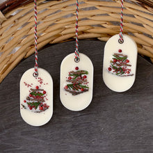 Load image into Gallery viewer, An elongated oval white wax tablet embellished with tiny dried branches, cranberry seeds, and red peppercorns resembles a Christmas tree. It features a red and green decorative rope threaded through a metal eyelet.