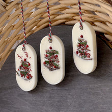 Load image into Gallery viewer, An elongated oval white wax tablet embellished with tiny dried branches, cranberry seeds, and red peppercorns resembles a Christmas tree. It features a red and green decorative rope threaded through a metal eyelet.