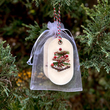 Load image into Gallery viewer, An elongated oval white wax tablet embellished with tiny dried branches, cranberry seeds, and red peppercorns resembles a Christmas tree. It features a red and green decorative rope threaded through a metal eyelet. Packaging: white organza bag.