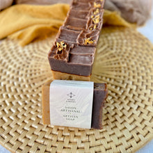 Load image into Gallery viewer, A row of handcrafted soap bars with a beige base and brown tops, adorned with orange peel. In front of the row is a single soap wrapped with a label from Woods & Mosses that reads Savon Artisanal and Artisan Soap.