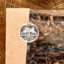 Load image into Gallery viewer, A close-up of a kraft gift box with a clear window, sealed with a round Hand Made sticker featuring vintage floral and ribbon illustrations. The packaging is filled with crinkle paper, highlighting the artisanal quality of the product inside.