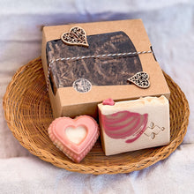 Load image into Gallery viewer, A natural soap gift set displayed on a woven tray. The set includes a heart-shaped pink soap and a rectangular soap with a pink swirl design, packed in a brown kraft box with a clear window and decorative heart accents. Elegant and handmade.