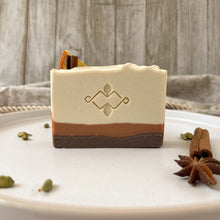 Load image into Gallery viewer, Front view of a handcrafted spiced orange soap bar with cream, caramel, and brown layers, stamped with an artisan logo and topped with dried orange and cinnamon for a festive, holiday-inspired look.