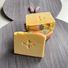 Load image into Gallery viewer, The square soap showcases a Woods and Mosses logo in the center. The soap body is yellow, while the top is a deeper shade of yellow, adorned with a small orange-green carrot decoration.