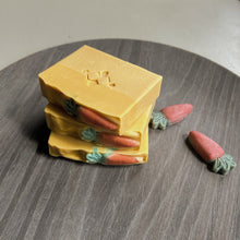 Load image into Gallery viewer, The square soap showcases a Woods and Mosses logo in the center. The soap body is yellow, while the top is a deeper shade of yellow, adorned with a small orange-green carrot decoration.