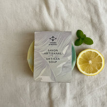 Load image into Gallery viewer, Round artisan soap partly wrapped in minimalist Woods and Mosses label showing lemon and mint tones.