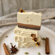 Load image into Gallery viewer, Stack of handmade spiced orange soap bars with layered cream, caramel, and brown tones, topped with dried orange peel and cinnamon sticks, showcasing a festive holiday design and stamped artisan logo.