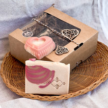 Load image into Gallery viewer, A natural soap gift set displayed on a woven tray. The set includes a heart-shaped pink soap and a rectangular soap with a pink swirl design, packed in a brown kraft box with a clear window and decorative heart accents. Elegant and handmade.