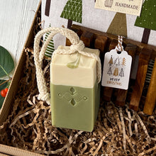 Load image into Gallery viewer, Two-tone green and cream Matcha Latte artisan soap bar with a braided natural cotton rope, placed in kraft paper filling beside a wooden soap tray and a holiday tag that reads Merry Christmas.