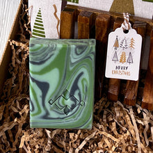 Load image into Gallery viewer, Green sage artisan soap bar with dark and light swirl pattern, nestled in kraft paper filling, shown next to a wooden soap dish and a festive Merry Christmas gift.