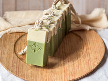 Load image into Gallery viewer, Line of vegan matcha soaps on a rope, arranged diagonally on a wooden surface. Natural skincare bars with embossed design and smooth top swirl.