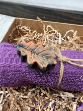 Load image into Gallery viewer, Autumn leaf–shaped orange‑black artisan soap resting on purple washcloth inside men’s self‑care gift set.