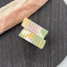 Load image into Gallery viewer, Top view of two handcrafted soap bars showing pastel stripe patterns in green, yellow, and pink, displayed on a round black wooden surface next to a peach cloth.
