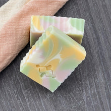 Load image into Gallery viewer, Two natural soap bars with marbled pastel swirls in green, yellow, and pink, angled together on a dark textured surface beside a soft woven cloth.