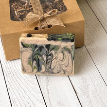 Load image into Gallery viewer, Handmade soap with green, black, and cream swirls, topped with a natural black accent, shown alongside eco-friendly packaging.