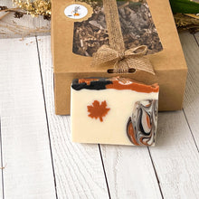 Load image into Gallery viewer, Natural artisan soap bar with a maple leaf design and swirling colors of orange, black, and cream, presented with eco-friendly packaging.