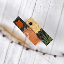 Load image into Gallery viewer, Two artisan soap bars with intricate swirling designs and a maple leaf motif, styled on a white wood background with a natural branch.