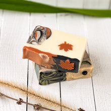 Load image into Gallery viewer, Stacked artisan soap bars with maple leaf motifs and natural swirls, styled with dried botanicals on a white wood background.