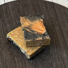 Load image into Gallery viewer, Two stacked handcrafted Autumn Ember Artisan Soap bars with a top layer of black and orange swirls and a bottom layer of natural exfoliating botanicals, displayed on a round dark wood tray. - vegan, handmade, moisturizing cleanser, eco-friendly gift idea by Woods and Mosses.