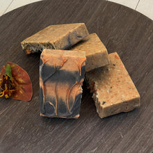 Load image into Gallery viewer, Two natural textured Autumn Ember Artisan Soap bars with a visible botanical mix and a black and orange swirl Autumn Ember Artisan Soap bar, accompanied by a dried flower, displayed on a round dark wood tray. - vegan, handmade, moisturizing cleanser, eco friendly gift idea.