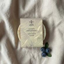 Charger l'image dans la galerie, Round artisan soap wrapped in a printed label reading “Savon Artisanal – Artisan Soap,” showing part of the bar’s natural texture beneath the band.