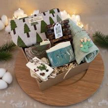 Load image into Gallery viewer, Vegan Christmas gift box with handmade natural soaps, wooden soap dish, green towel, and holiday pouch in eco-friendly packaging, displayed with pine branches and festive lights on a wooden tray.