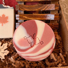 Load image into Gallery viewer, A round soap bar featuring bold red, pale rose and white swirls, with the company's logo prominently displayed at its center.