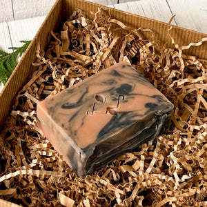 Handcrafted cold process soap bar with a black and terracotta swirl pattern, featuring a stamped geometric logo on top, nestled in kraft paper shred. Part of a natural men's skincare gift set for winter.