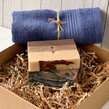 Load image into Gallery viewer, Stacked artisan soap bars in a kraft box. One layered with earthy tones and a real leaf embed, the other with black and terracotta swirls, paired with a rolled navy washcloth, part of a holiday gift set for men’s natural skincare and winter wellness.