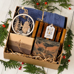 Men’s holiday gift box featuring two artisan soap bars, one marbled, one layered. A wooden soap tray, navy cotton towel, laser-cut Santa ornament, and a Merry Christmas tag, all nestled in eco-friendly kraft packaging.