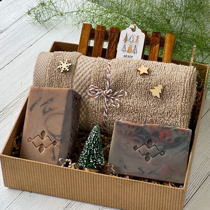 Gift box with two artisan soap bars in earthy tones, a wooden soap dish, and a folded beige textured towel decorated with small wooden holiday shapes, arranged with kraft filler and a miniature snow-dusted Christmas tree for a festive presentation.