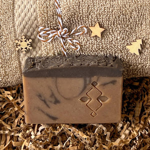 Handcrafted brown soap bar with a dark textured top and subtle black swirls, embossed with a geometric design, displayed on kraft paper filler in front of a folded beige towel decorated with small wooden holiday shapes.