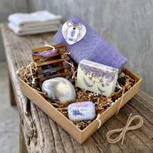 Load image into Gallery viewer, A kraft gift box filled with multicolor raffia paper, containing a white-purple square and round soap bars, a wooden soap dish, a soft purple cotton face cloth, a decorative lavender-pink tin filled with dried lavender buds, and a paper tag.
