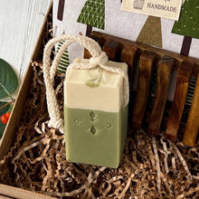 Load image into Gallery viewer, Two-tone green and cream Matcha Latte artisan soap bar with a braided natural cotton rope, placed in kraft paper filling beside a wooden soap tray.