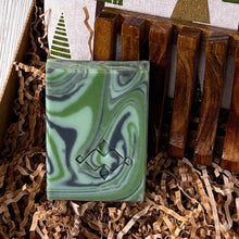 Load image into Gallery viewer, Green sage artisan soap bar with dark and light swirl pattern, nestled in kraft paper filling, shown next to a wooden soap dish.