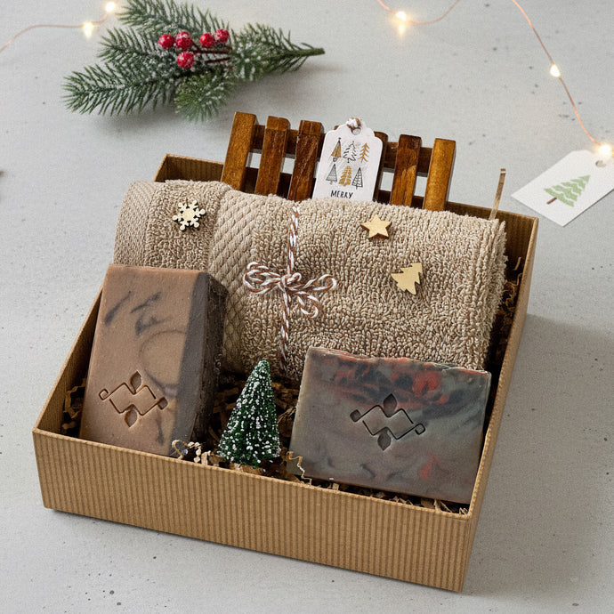 Gift box with two artisan soap bars in earthy tones, a wooden soap dish, and a folded beige textured towel decorated with small wooden holiday shapes, arranged with kraft filler and a miniature snow-dusted Christmas tree for a festive presentation.