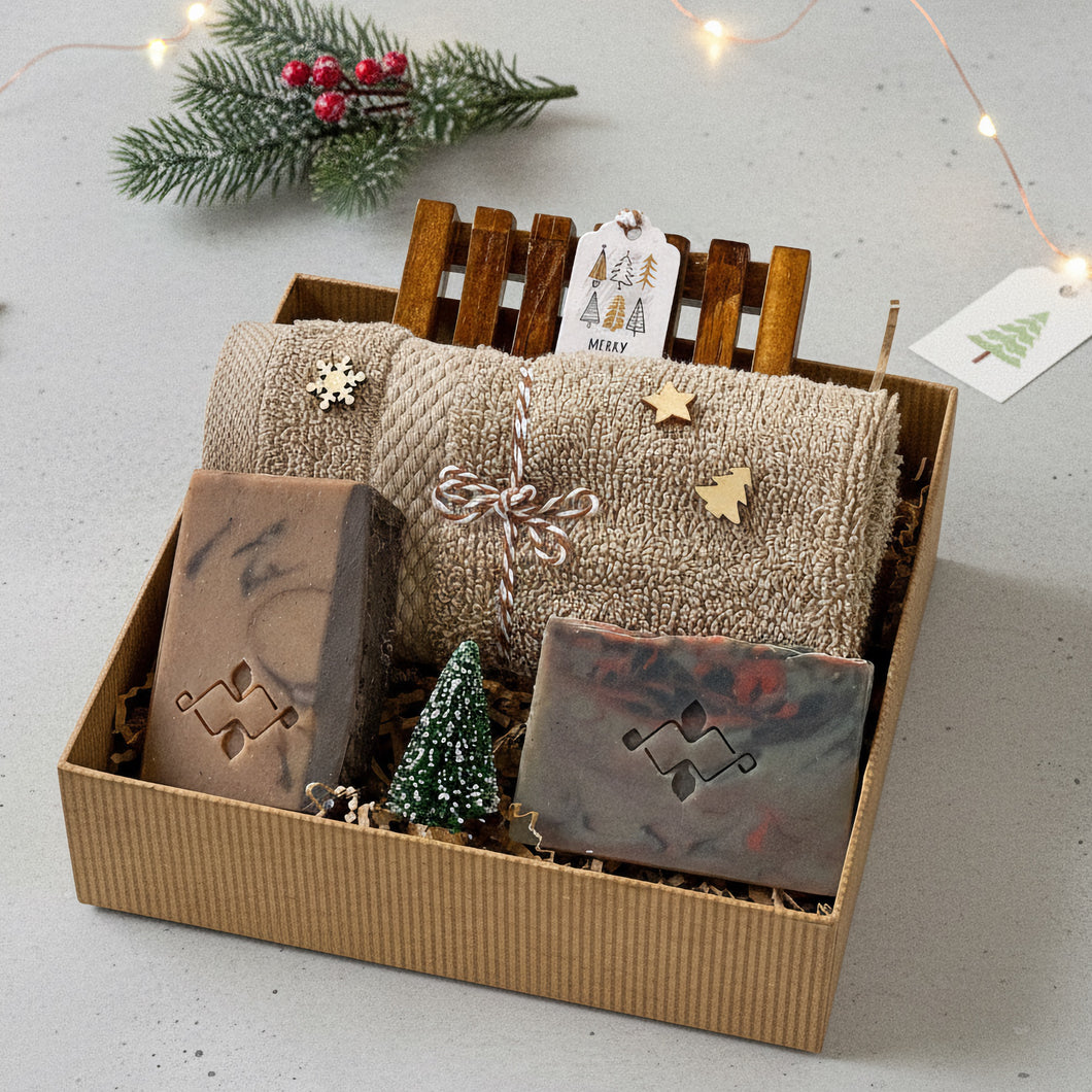Gift box with two artisan soap bars in earthy tones, a wooden soap dish, and a folded beige textured towel decorated with small wooden holiday shapes, arranged with kraft filler and a miniature snow-dusted Christmas tree for a festive presentation.