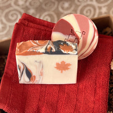 Load image into Gallery viewer, Beige soap bar featuring an orange maple leaf design and a round soap bar featuring bold red, pale rose and white swirls, both placed on a red cotton facial towel.