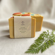Load image into Gallery viewer, Wrapped artisan soap bars with visible label and carrot-detail bars behind. Handmade Skincare, Montreal, Canada.