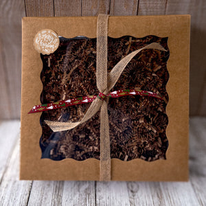 Eco-friendly kraft gift box with a scalloped window, filled with crinkle paper and tied with natural twine and a red festive ribbon, featuring a round Merry Christmas sticker in the corner.