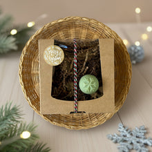 Load image into Gallery viewer, A kraft box with a transparent lid filled with brownish soap, tied with a festive white, red, and green rope and adorned with a Merry Christmas sticker. A round pale green mini soap featuring a delicate snowflake print on the top.