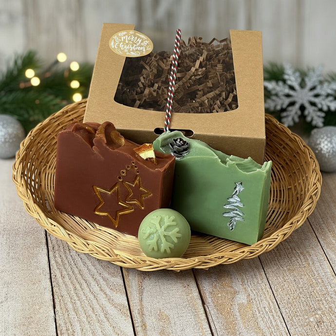 A kraft box with a transparent lid filled with brownish soap, tied with a festive white, red, and green rope and adorned with a 'Merry Christmas' sticker. Beside the box are brownish square soap, green square soap, and a round pale green mini soap.