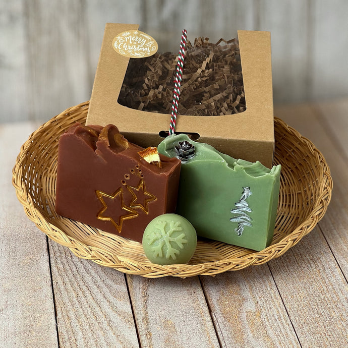 A kraft box with a transparent lid filled with brownish soap, tied with a festive white, red, and green rope and adorned with a 'Merry Christmas' sticker. Beside the box are brownish square soap, green square soap, and a round pale green mini soap.
