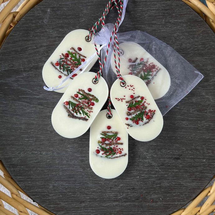 An elongated oval white wax tablet embellished with tiny dried branches, cranberry seeds, and red peppercorns resembles a Christmas tree. It features a red and green decorative rope threaded through a metal eyelet. Packaging: white organza bag.